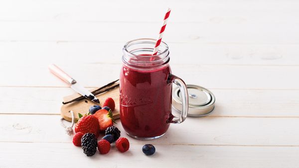 Smoothie vermelho num copo com bagas vermelhas, frutas e baunilha na mesa.