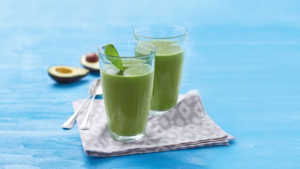 Two green smoothies in glasses arranged together with avocado halves on table.