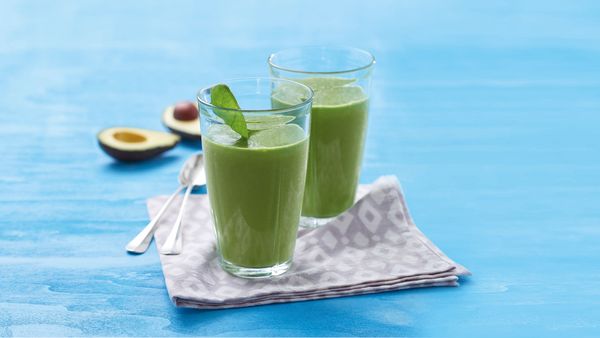 Two green smoothies in glasses arranged together with avocado halves on table.