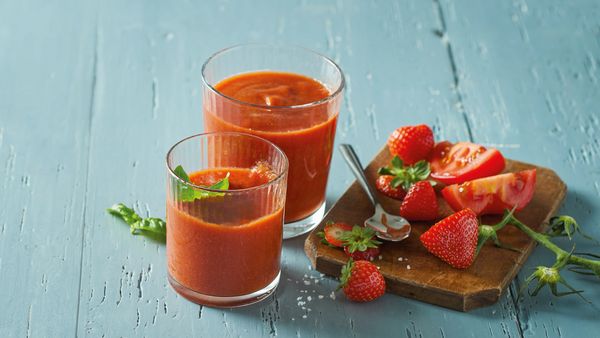 Two red smoothies in glasses arranged together with strawberries and tomatoes on table.