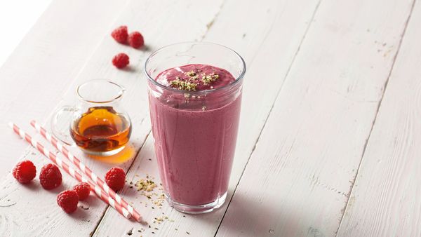 Pink smoothie in a glass arranged together with a small jar of maple syrup and a couple of raspberries.