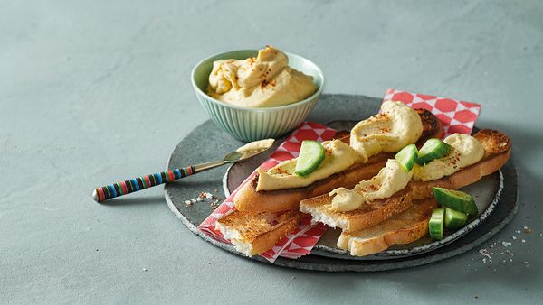 Small bowl filled with chickpea hummus arranged together with slices of toast spread with hummus on a plate.