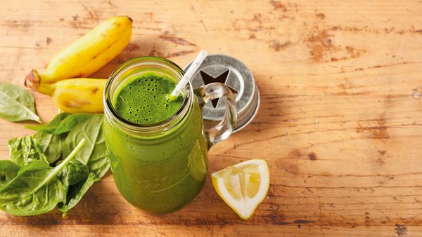 Smoothie verde într-un pahar aranjat împreună cu frunze de spanac, banane și o lămâie pe o masă.