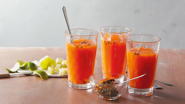 Drei orangefarbene Smoothies in Gläsern, angerichtet mit Apfel- und Limettenscheiben auf einem Tisch.