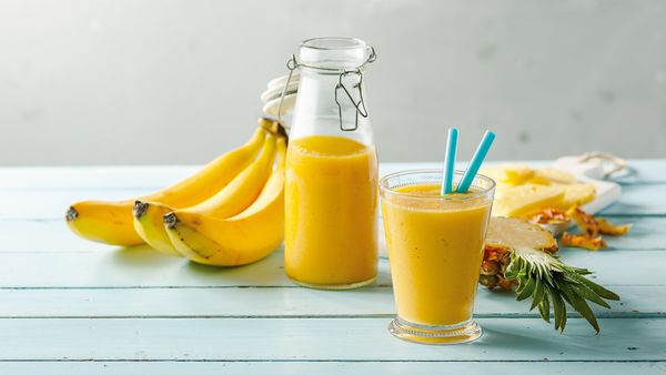 Bicchiere pieno di frullato giallo e un boccale per succo sistemati su un tavolo insieme ad alcune banane, fette di mango e mezzo ananas.