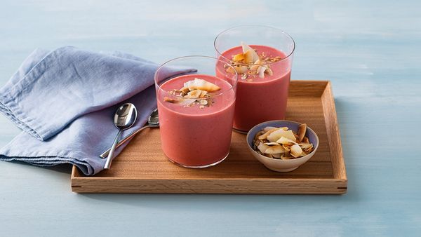 Două shake-uri roșiatice în pahare aranjate împreună cu un bol mic cu semințe de floarea soarelui și fulgi din nucă de cocos pe o tavă.