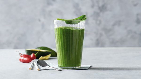 Grüner Smoothie in einem Glas, angerichtet mit einer Avocadohälfte und einer Chilischote.