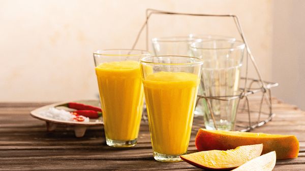 Dos batidos amarillos hechos de mangos rellenos en vasos y dispuestos junto con rodajas de mango.