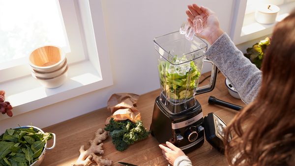 Mulher a adicionar cubos de gelo e legumes num jarro de um liquidificador VitaBoost.