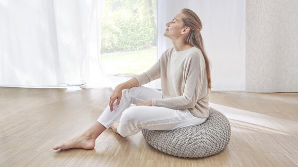 A woman sits on a pouf in the living room and takes a deep breath.