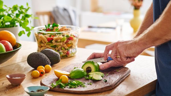 Eine Person schneidet an einer Kücheninsel auf einem Holzbrett Avocados in Scheiben. Im Hintergrund steht eine Schüssel mit Salat.