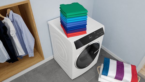 Billede nedefra af sammenlagte håndklæder i forskellige farver stablet oven på en vaske-/tørremaskine, der står ved et åbent garderobeskab og en vasketøjskurv. 