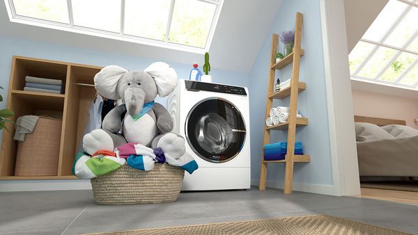 Im Vordergrund sitzt ein großer Plüschelefant in einem Wäschekorb. Im Hintergrund steht ein Waschtrockner in einem hellen Wäscheraum. 
