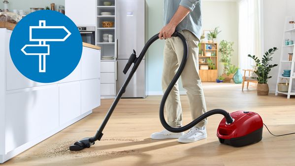 Ein Mann saugt mit einem roten kabelgebundenen Bosch Staubsauger mit Beutel  auf Hartboden in einem modernen Wohnraum mit offener Küche.