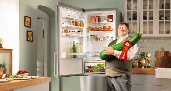 Мъж, който държи гигантска тиквичка в ръцете си и стои в кухня до изключително голям хладилник с фризер.