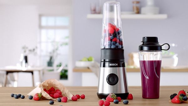 Mini blender Bosch VitaPower Serie 2 cu fructe roșii și o sticlă To-Go umplută cu smoothie pe un raft de bucătărie.