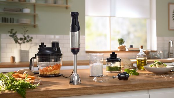 Hand blender ErgoMaster Series 6 standing on a worktop next to the attachments cube cutter, chopper, whisk and ingredients.