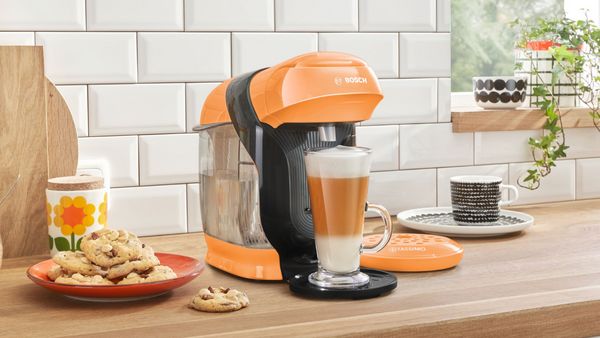 TASSIMO STYLE in the colour peach brewing a Latte Macchiato on a kitchen worktop and a plate of cookies next to it.