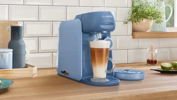 TASSIMO FINESSE in the colour lupine blue brewing a Latte Macchiato on a kitchen worktop and a plate of pancakes next to it.