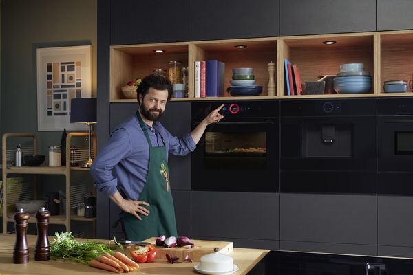 En mann på et kjøkken med et forkle med gulrottrykk. Grønnsaker inni en Bosch Serie 8-ovn, fingrene hans berører berøringsskjermen.