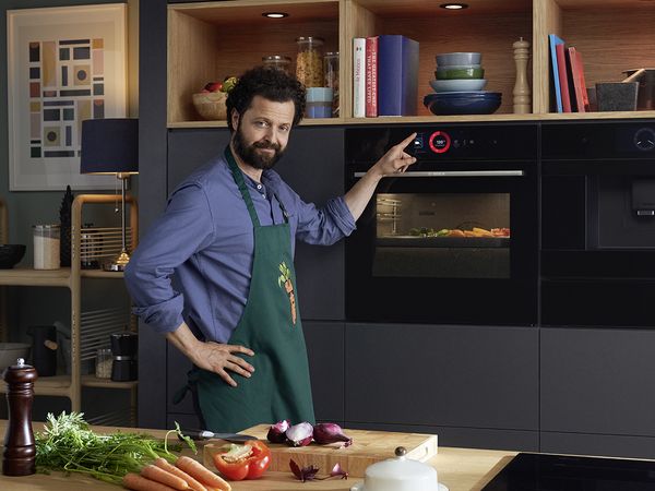 Muž v kuchyni s oblečenou zásterou s potlačou mrkiev. Zelenina je vložená vo vnútri rúry na pečenie Bosch série 8, pričom jeho prst stláča dotykový displej.