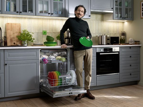 A man is standing in front of a dishwasher. He holds a dried plate in his hand and smiles.