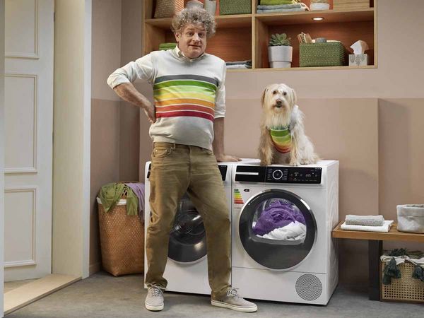 En mann som står i et vaskerom med en Bosch varmepumpetørketrommel. Smiler fordi den energieffektive varmepumpetørketrommelen sparer energi takket være den mest effektive energimerkingen som også er tegnet på genseren.