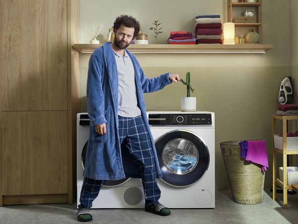 A cactus with arms stands on a washing machine with the sustainable i-DOS automatic dosing system. A man in a bathrobe gives the cactus a fist-bump.