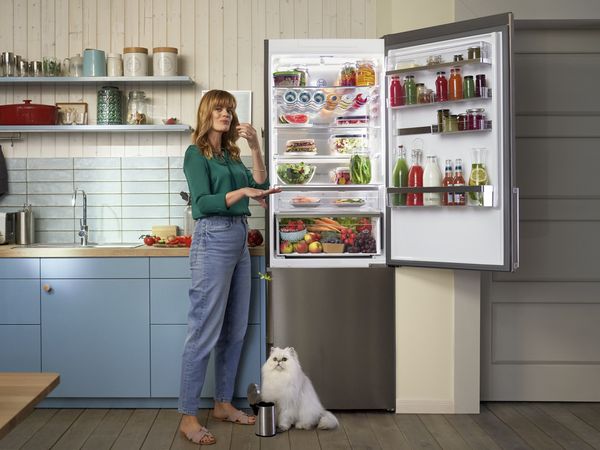 Eine Frau steht in einer Küche neben einem offen stehenden Kühlschrank, welcher voller frischer Lebensmittel ist. Sie isst gerade etwas frisches aus dem Kühlschrank, während eine weiße Katze auf dem Boden sitzt.