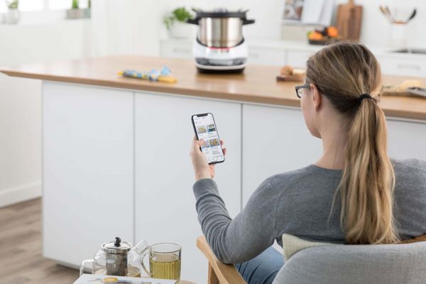 Una donna davanti a Cookit, con uno smartphone su cui si vede l’app Home Connect aperta. 