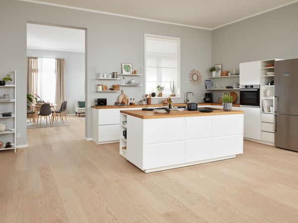 Una generosa cocina blanca, totalmente equipada con isla de cocción y paso a la sala-comedor. El suelo es de madera clara y las grandes ventanas dejan entrar mucha luz.