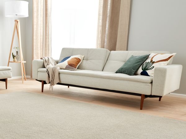 Una luminosa sala con sofá y sillones Sobre el suelo de madera hay una alfombra rectangular La luz entra por una gran ventana situada detrás del sofá