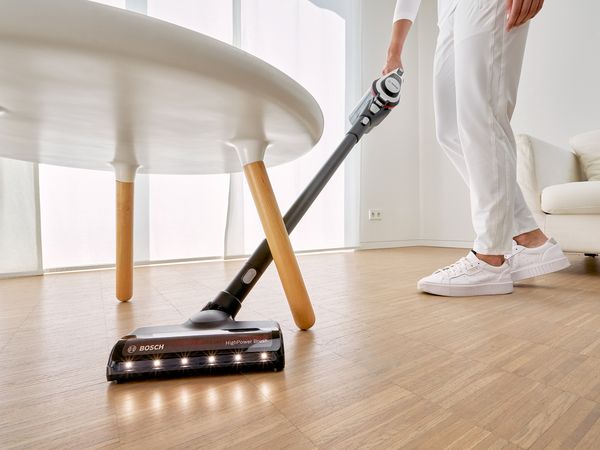 Someone is vacuuming a wooden floor with the cordless vacuum cleaner Unlimited 8. The vacuum cleaner with lights at the front of the nozzle manoeuvres easily under a table.
