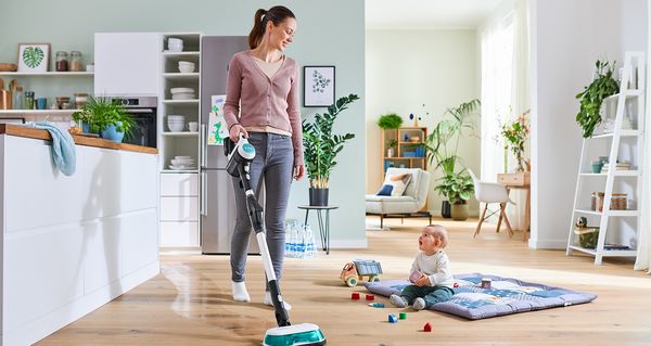 Une jeune maman est en train de laver le sol de la cuisine avec l'Unlimited 7 ProHygiène Aqua. À côté d'elle, un jeune enfant joue avec des petits jouets sur une couverture posée sur le sol en bois.