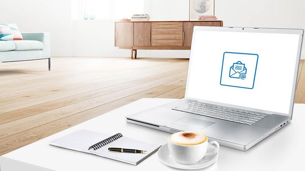 Ein aufgeklappter Laptop, ein Ringbuch und Stift sowie eine Tasse Cappuccino mit Milchschaum stehen auf einem weißen Tisch in einem modernen Wohnraum.