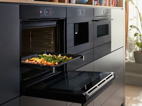 Elegantna kuhinja z Bosch vgradno pečico serije 8 s funkcijo Air Fry in grelnim predalom. Različna zelenjava, kuhana v pečici. Več zelenjave na deski za rezanje na pultu.