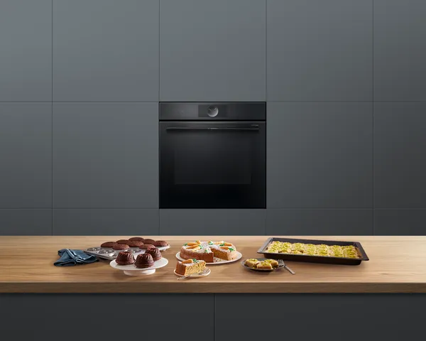 Four électrique Série 8 de Bosch dans une cuisine. Sur un comptoir devant lui se trouvent divers produits de boulangerie tels qu'un gâteau aux carottes et des muffins.