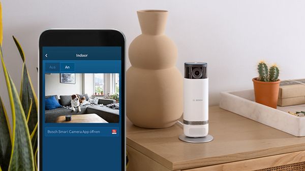 Die Bosch Smart Home Innenkamera, auf einem kleinen weißen Regal und ein Smartphone auf dem die Liveaufnahme der Kamera abgebildet ist.