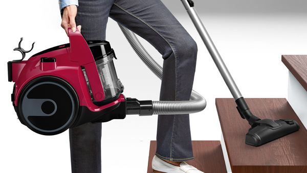 En person bærer en rød cylinder fra en Bosch støvsuger uden pose, mens trappen rengøres.