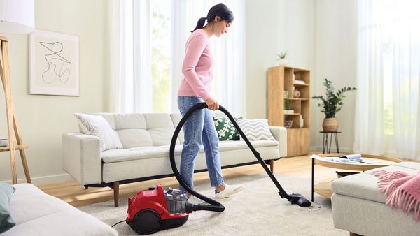 A woman uses a red Bosch bagless vacuum to clean a bright, airy living room.