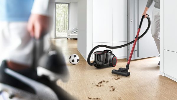 Schmutz wird mit einem schwarz-roten ProPower Staubsauger ohne Beutel von Bosch von einem Hartboden gesaugt. Auf dem Boden liegt ein Fußball und eine Person trägt Fußballschuhe durch den Raum.