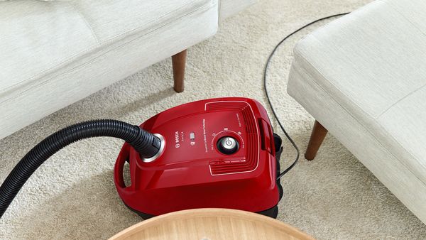 Kompaktowy, czerwony workowy odkurzacz cylindryczny marki Bosch manewruje pomiędzy dwiema sofami i stołem.