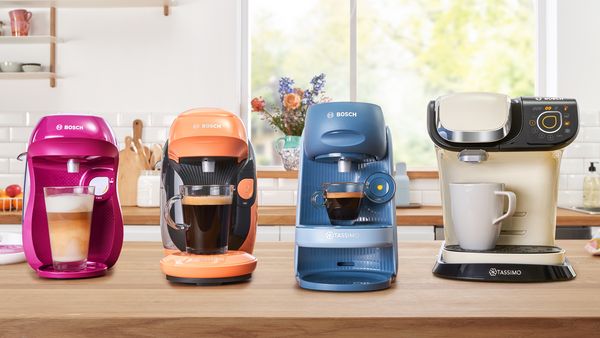 4 modely kávovarů TASSIMO na kuchyňské desce a 4 různé kávové speciality.