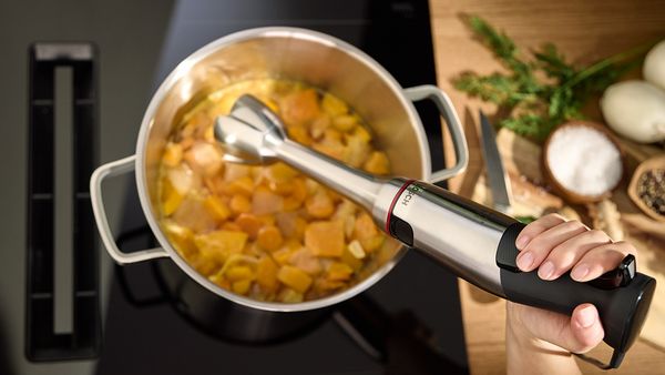 Ręka trzymająca blender zanurzeniowy nad garnkiem ze świeżo ugotowanymi warzywami, gotowy do przygotowania gładkiej zupy.