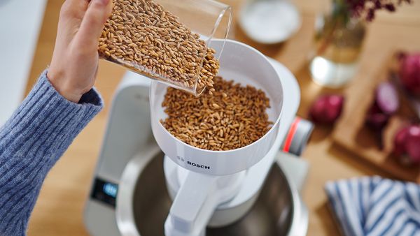 Eine Person gibt Getreide aus einem Glas in die Getreidemühle einer Multifunktions-Küchenmaschine.