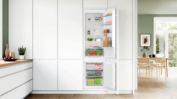 Een van de kastdeuren in een witte keuken die op de eetruimte uitloopt, is blijven open staan en laat de goed gevulde ingebouwde XL koel-vriescombinatie erachter zien.