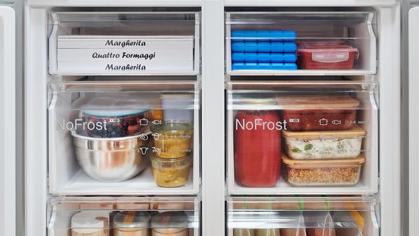 Detailní pohled na zásuvky mrazničky s logem No Frost ukazuje, že jsou plné nádob se zbytky jídla a potravin bez ledu.