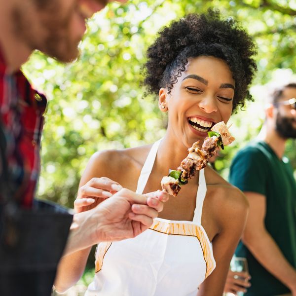 A woman is being handed a skewer with meat and vegetables on it, a man with sunglasses stands behind her.