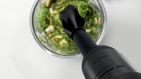 A close up image of a handheld food processor making pesto in a glass bowl.