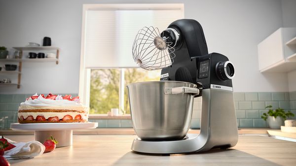 Bosch stand mixer with a whisk next to a cake and whipped cream.